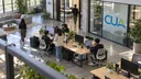 Coworkers share computer stations in an open studio with the Cua logo on a glass partition.