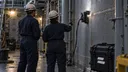 Two naval maintenance workers watch an inspection crawler on a ship’s steel bulkhead beside a Gecko Robotics equipment case.