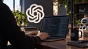 A person types code on a laptop beside an OpenAI logo on the wall and a mug, with a small robot nearby.