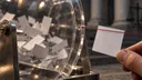 Blank raffle slips fill a drum while one is held apart, evoking Elon Musk’s PAC giveaway and class-action settlement.