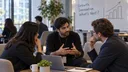 AI-generated illustration of three people talking around a laptop, with a whiteboard behind them showing handwritten words and an upward bar chart.